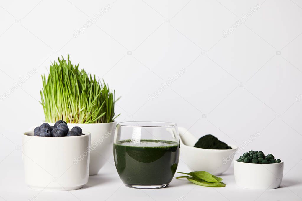 fresh smoothie from spirulina, cups with spirulina grass and blueberries, leaves, bowls with spirulina powder and spirulina pills on grey background 