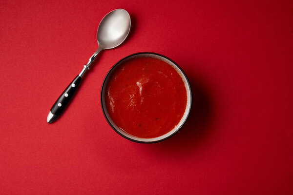 top view of tomato soup in plate and spoon on red surface