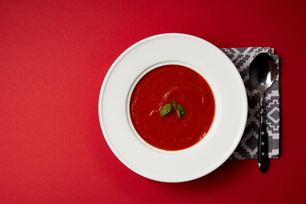 top view of tasty tomato soup in plate with spoon and napkin on red table