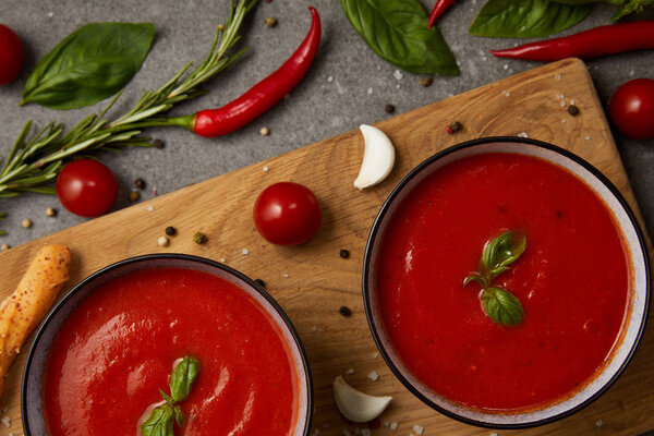 top view of tasty tomato soup in plates on cutting board with vegetables on grey table