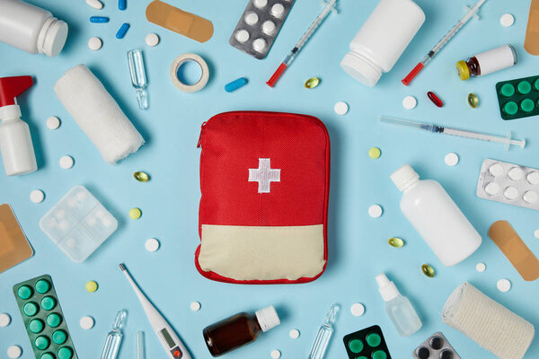 top view of red first aid kit bag on blue surface surrounded with different medicines