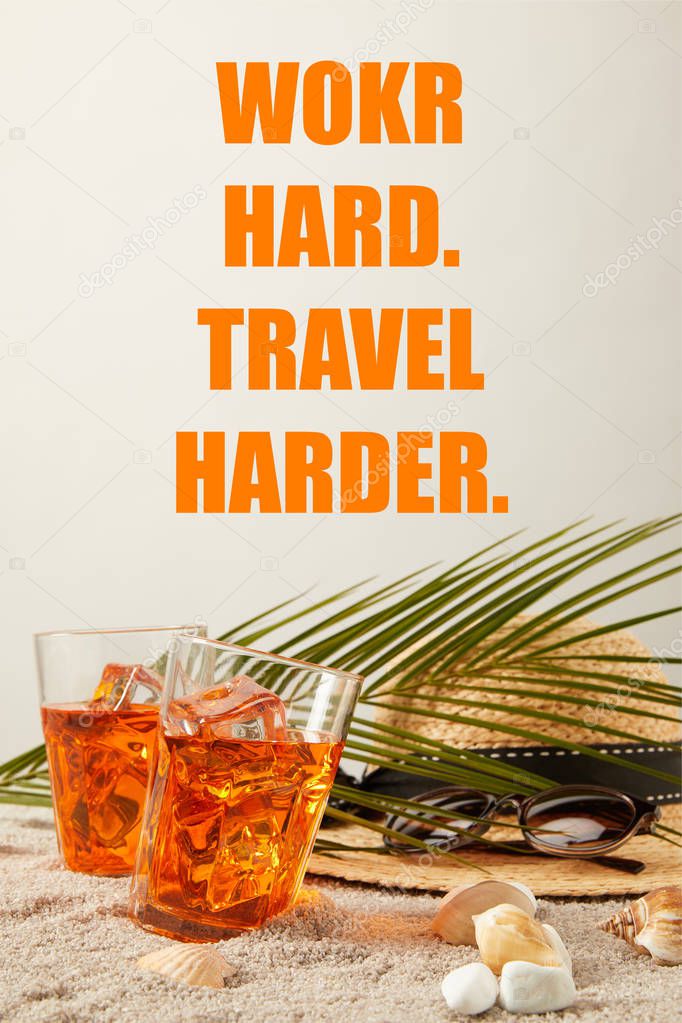 close up view of straw hat, cocktails, seashells, sunglasses and palm leaf on sand on grey backdrop, work hard travel harder inscription