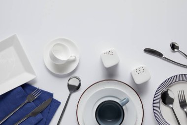 top view of various ceramic plates, saltcellar, pepper caster, cup, kitchen towel, forks, spoons and knives on white table  clipart