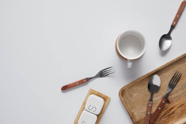 top view of cup, spoons, forks, wooden tray, saltcellar and pepper caster on white table  clipart