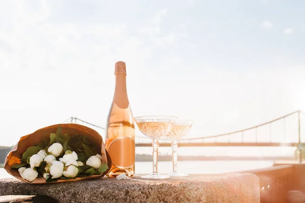 stock image close-up view of beautiful bouquet of flowers, two glasses and bottle of champagne near river at sunset 