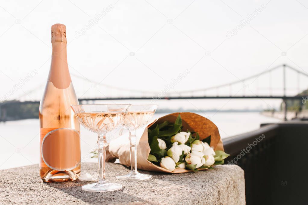 beautiful bouquet of flowers, glasses and bottle of champagne near river at sunset 