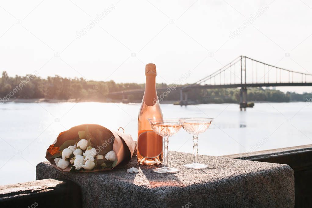 bottle of champagne, two glasses and beautiful bouquet of flowers on embankment at sunset