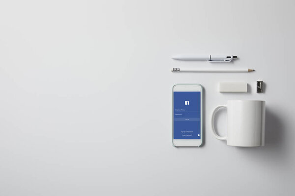 top view of smartphone with facebook app and various supplies on white tabletop
