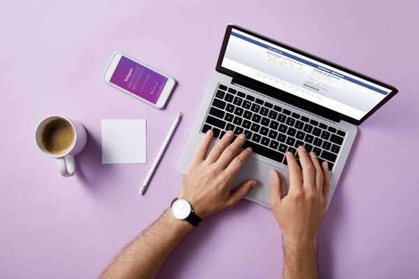 cropped shot of man using laptop with facebook website on screen and instagram app in smartphone at workplace on pink surface