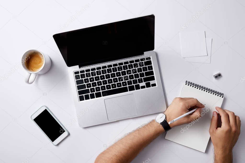 cropped shot of man writing in notebook at workplace with gadgets and coffee cup on white surface for mockup