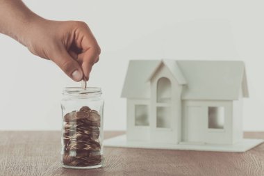 cropped image of man putting coin into glass jar with small house on table, saving concept clipart