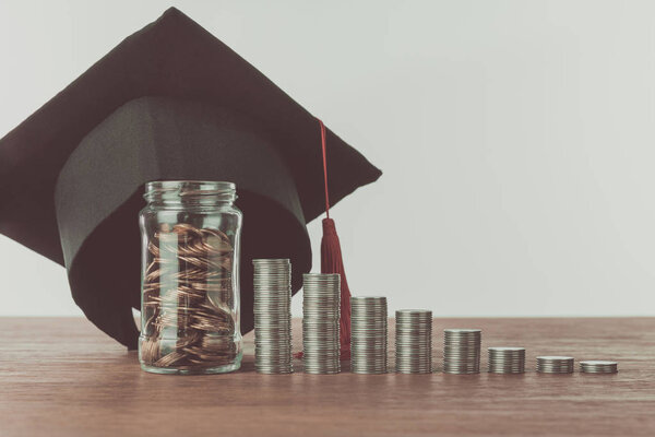 stacks of coins, jar with coins and graduation cap on wooden table, saving concept