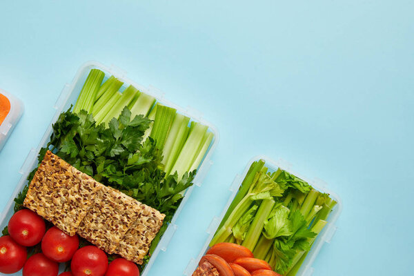 top view of arrangement of food containers with fresh healthy vegetables and cookies with seeds isolated on blue