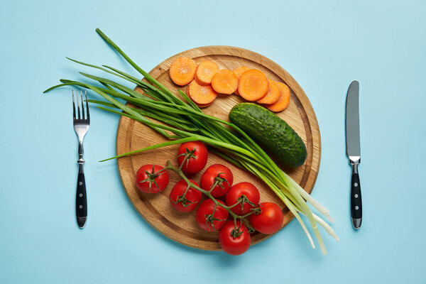 top view of cutlery and assorted fresh vegetables on wooden cutting board isolated on blue