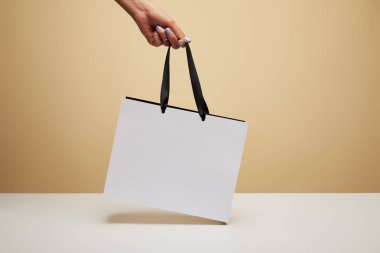cropped image of woman holding white shopping bag above white table isolated on beige clipart