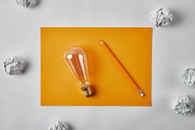 top view of incandescent lamp on blank yellow paper with pencil surrounded with crumpled papers on white surface clipart