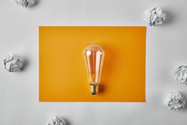 top view of incandescent lamp on blank yellow paper surrounded with crumpled papers on white surface clipart