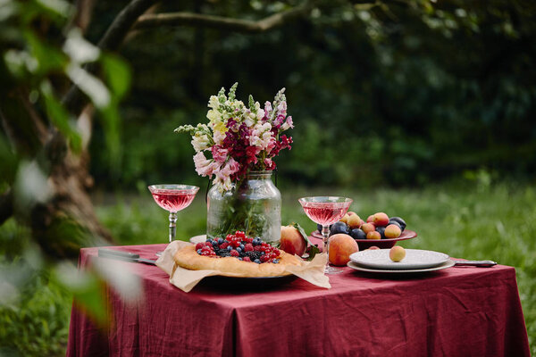 bouquet of flowers in glass jar, fruits and wineglasses on table in garden