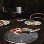 Slice of pizza with server on metal tray and wooden table with plates of lasagne and syrniki