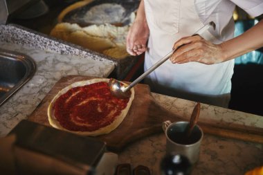 cropped shot of chef pouring ketchup onto pizza dough at restaurant kitchen clipart