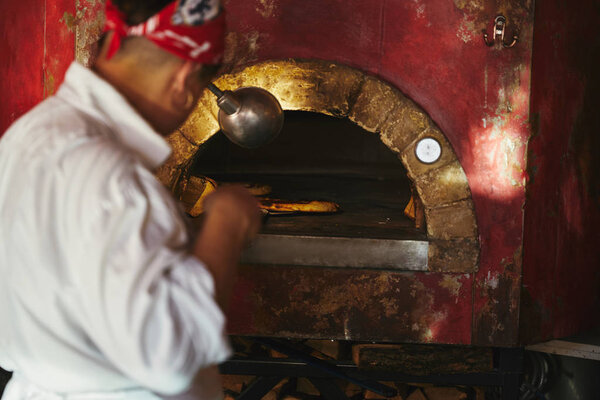 cropped shot of chef taking pizza from stone oven at restaurant kitchen