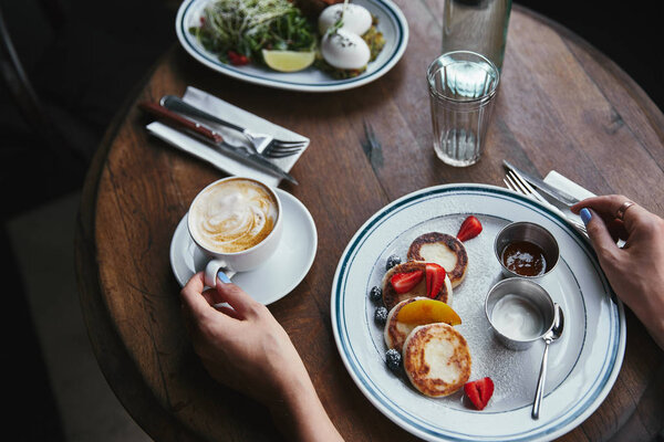 cropped shot of woman with cheese pancakes and coffee on wooden table at restaurant