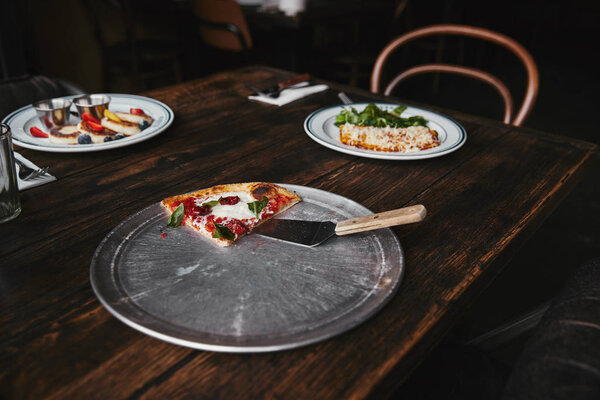 slice of tasty pizza with server on metal tray and wooden table with plates of lasagne and curd pancakes