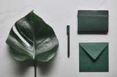 Zelená obálka a zápisník s perem na bílém mramorovém pozadí s monstera list