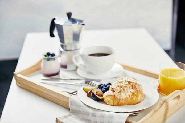 close up view of croissant with fresh blueberries and plum pieces, glass of juice, cup of coffee and yogurt for breakfast on wooden tray on white tabletop