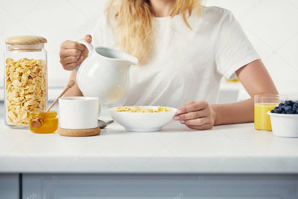 partial view of woman with jug of milk sitting at table with bowl of corn flakes and glass of juice for breakfast at home