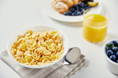 close up view of corn flakes in bowl, glass of juice and croissant with blueberries and plum pieces for breakfast on white tabletop clipart