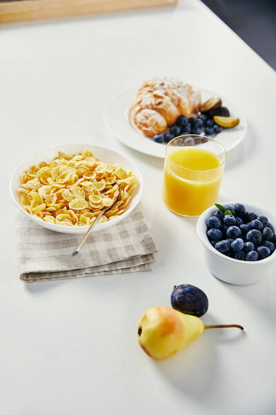 закрыть вид на кукурузные хлопья в миске, стакан сока и круассан с черникой и кусочками сливы на завтрак на белом столе

