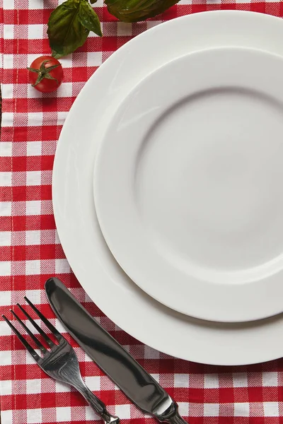市松模様のテーブル クロスで空の白い皿 カトラリー トマトとバジルのトップ ビュー — ストック写真