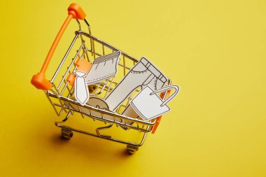 close up view of little shopping cart with clothes made of paper on yellow background