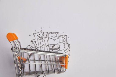top view of shopping cart with little goods made of paper on grey background clipart