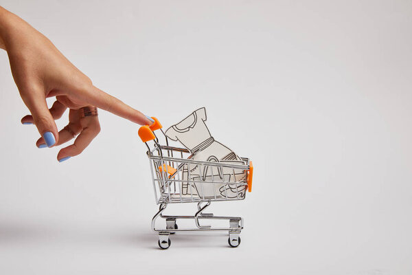 cropped shot of female hand and little shopping cart with paper clothes on grey background