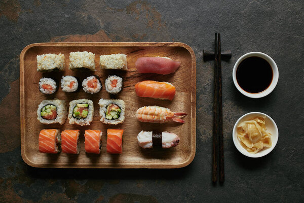 top view of assorted sushi set on wooden plate, chopsticks, ginger and soya sauce in bowls on dark tabletop