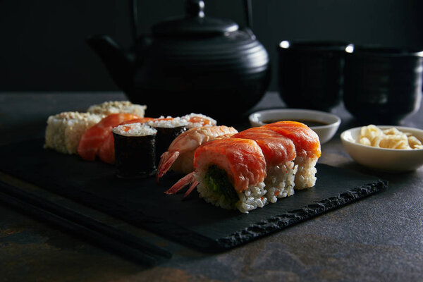 food composition with sushi set, ginger and soya sauce in bowls, teapot and cups with tea on dark surface
