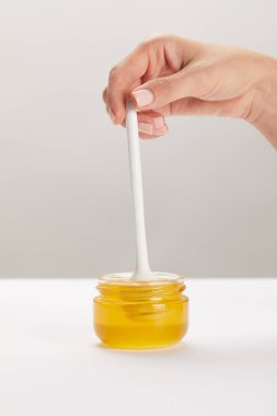 partial view of woman holding wooden honey deeper with glass jar full of honey on white surface clipart