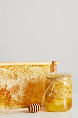 close up view of wooden honey deeper, glass jar with honey and stack of beeswax on white tabletop on grey background  clipart