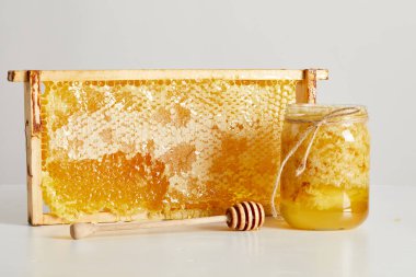 close up view of wooden honey deeper, glass jar with honey and stack of beeswax on white tabletop clipart
