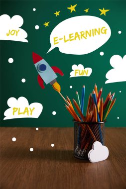 colorful pencils, rocket, cloud sign on wooden table with chalkboard on background with play, joy, fun and e-learning words clipart
