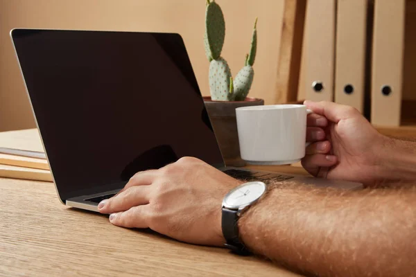 空白の画面を持つノート パソコンを使用して テーブルでコーヒー カップを保持している男のトリミングされた画像  — 無料ストックフォト
