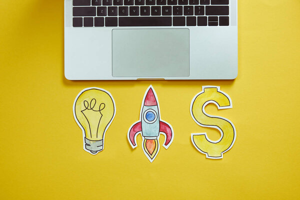 top view of laptop, paper lightbulb, paper rocket and dollar sign on yellow surface