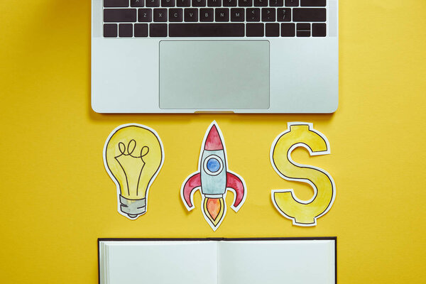 повышенный вид ноутбука, бумажной лампочки, бумажной ракеты, знака доллара и ноутбука на желтой поверхности
