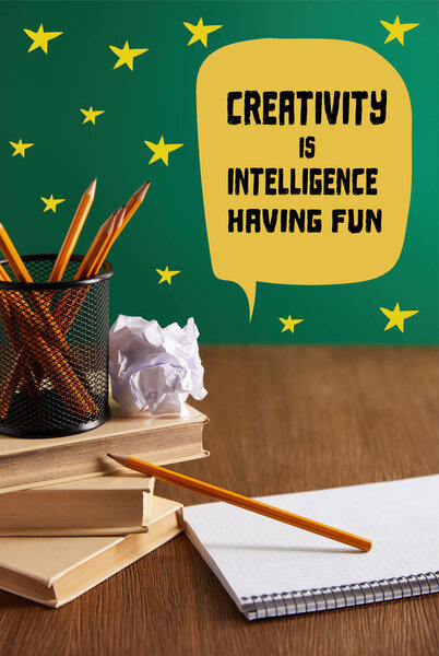 books, copybook, crumpled paper and pencils on wooden table with "creativity is intelligence, having fun" lettering