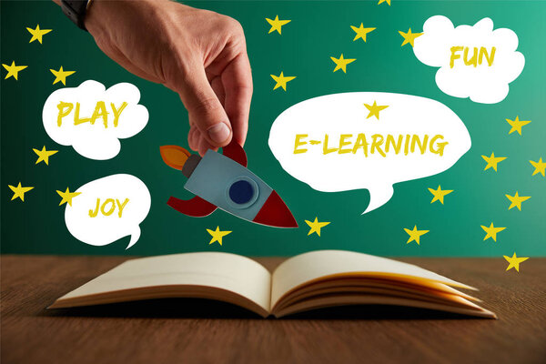 cropped view of teacher holding rocket over open book with play, joy, fun and e-learning words