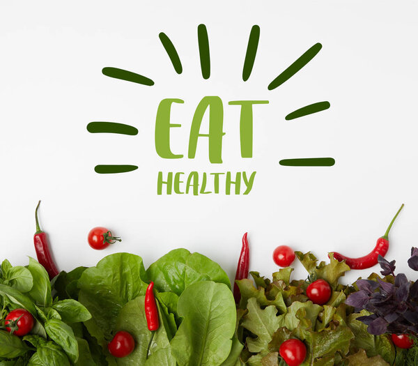 top view of fresh various leaf vegetables with tomatoes and peppers on white surface with "eat healthy" lettering
