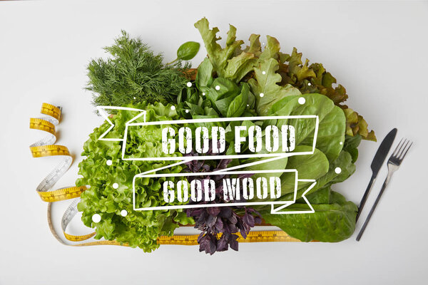top view of various ripe vegetables in box with cutlery and measuring tape on white surface with "good food - good mood" inspiration