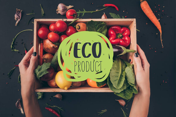 cropped image of woman holding wooden box with vegetables above gray table with "eco product" lettering